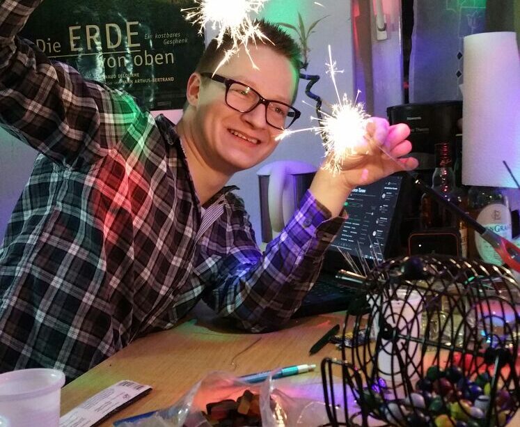 Kevin hält zwei brennende Wunderkerzen während der Silvesterfeier, es gibt buntes Licht und auf einem Tisch steht ein Bingo-Spiel.